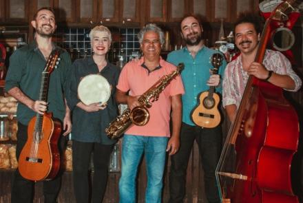 Grupo Choro do Jura posa para câmera enquanto seguram instrumentos. Ao todo são cinco pessoas, sendo quatro homens e uma mulher. O primeiro segura um violão, a segunda segura um pandeiro. O terceiro integrante da banda segura um saxofone, já o quarto musico tem um cavaquinho. Por fim, o último homem está com um violoncelo. 