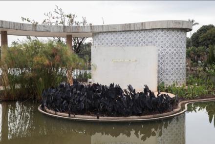 A imagem apresenta um laguinho e um jardim da Casa do Baile. É possível ver plantas roxas e pequenas árvores, além da construção da Casa. 