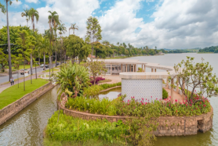 Fachada lateral da casa do baile, cercada pelo jardim florido e com árvores. Ao fundo está a Lagoa da Pampulha