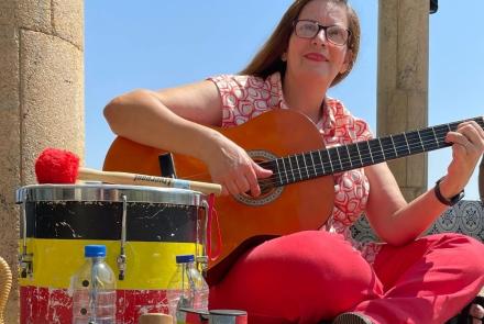 Alguns instrumentos musicais coloridos estão apoiados no chã, como um tambor. Uma mulher de roupa rosa está senta um pouco mais ao fundo, ela segura um violão e olha para câmera. Ela é branca, possui cabelos vermelhos e usa óculos.