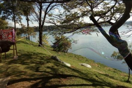 Mãos negras seguram dois palitos amarelos, usados para fazer uma grande bola de sabão. A imagem apresenta um gamado e árvores, em um dia ensolarado. Ao fundo, parte da Lagoa da Pampulha. 