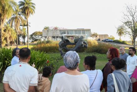 Grupo de pessoas de costas para a câmera observam ao jardim e fachada do Museu de Arte da Pampulha, localizado ao fundo da imagem.