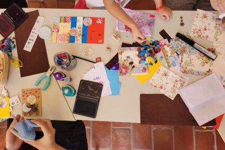 Mesa com diversos recortes coloridos e ferramentas para artesanato, como alicate, régua, tesoura, cola. Mãos de duas pessoas brancas aparecem manuseando os materiais. 