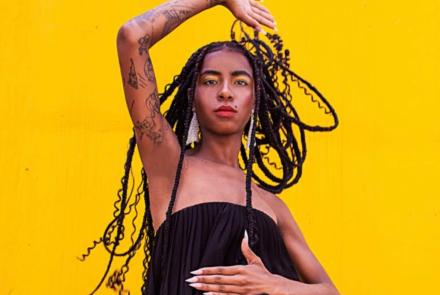 Mulher negra posando para foto com o braço direito para cima e o esquerdo no corpo. Ela está com uma roupa preta e com cabelos trançados esvoaçantes. Ela está em frente a um fundo amarelo.