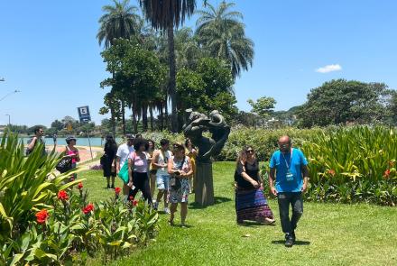 Grupo de pessoas caminha pelos jardins do Museu de Arte da Pampulha. O dia está ensolarado, a grama verde e o céu azul. Há coqueiros ao fundo