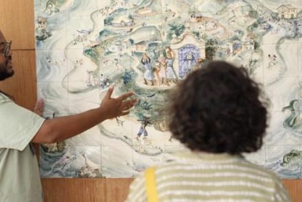 Uma mulher, que está de costas para câmera, observa um quadro feito de azulejos coloridos, enquanto um homem negro faz a explicação da obra. Ela possui cabelos curtos e está usando roupa verde e branca. Ele possui cabelos longos e trançados. 