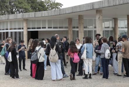 Um Grupo de pessoas aguardam em pé na porta da Casa do Baile. Elas usam mochilas. 