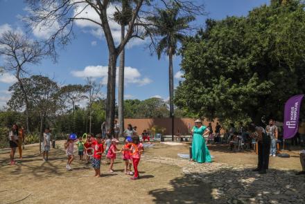 Grupo de crianças brincando no gramado de um jardim. eles estão acompanhados por alguns adultos ao redor. Uma mulher de vestido verde fotografa. 