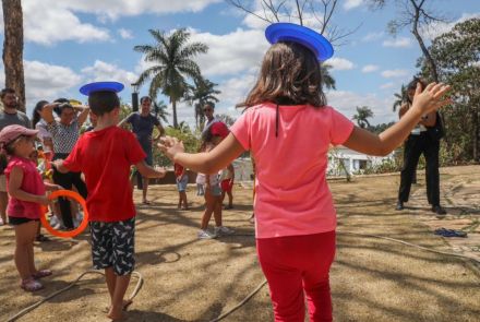 Duas crianças de roupas vermelhas, equilibram um pratinho azul na cabeça. Elas estão em um gramado, Outras crianças aparecem ao fundo