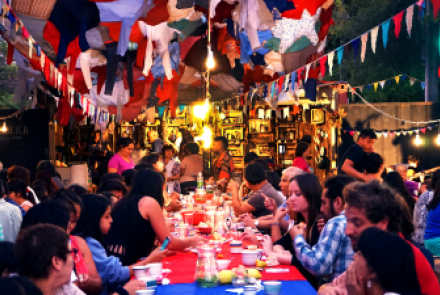 Na foto, pessoas compartilham comidas em uma mesa comunitária