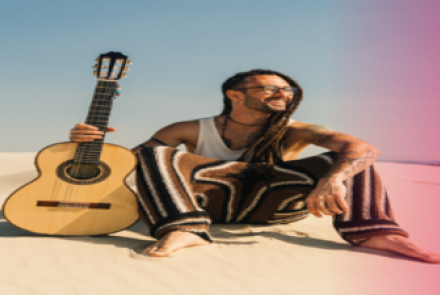 Foto mostra um homem sentado na praia segurando um violão