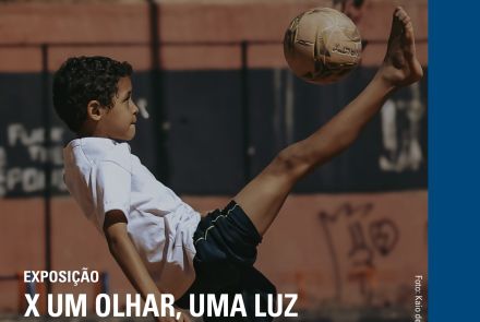Um menino joga futebol em um campo de terra. Ele está chutando a bola com o pé levantado, usando camiseta branca e shorts escuros. Ao fundo, há um muro com tijolos e pichações. Na parte de cima, está escrito: “04 a 30 de novembro de 2025 – Exposição X Um Olhar, Uma Luz – Secretaria Municipal de Educação de Belo Horizonte.” Abaixo, diz: “MM Gerdau – Museu das Minas e do Metal, Praça da Liberdade, número 680, Prédio Rosa, Belo Horizonte/MG.”
