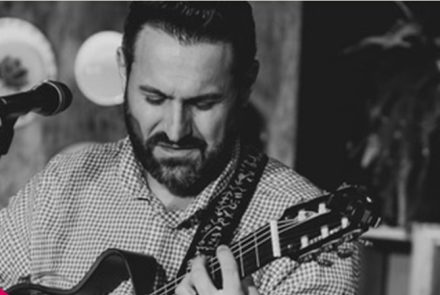 Imagem preta e branca, com um homem tocando um instrumento com um microfone na sua frente