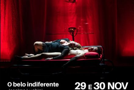 Espetáculo: "O Belo Indiferente" - Cena teatral iluminada por luz vermelha intensa. Uma mulher está deitada sobre um homem em uma cama metálica, em um momento de forte carga dramática. Ao fundo, cortinas vermelhas e uma escultura pendurada compõem o cenário.