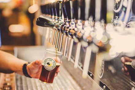 Imagem de varias torneiras de chopp, e uma pessoa enchendo o copo de chopp