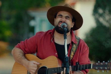 Um homem toca violão e canta ao ar livre. Ele usa chapéu marrom, camisa vermelha e toca diante de um microfone. Atrás dele há casas antigas coloridas e árvores.