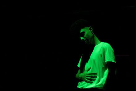 A foto mostra uma pessoa em um ambiente escuro, iluminada por uma luz verde. Ela está usando uma camiseta clara e aparece com uma das mãos sobre o abdômen. O fundo é totalmente preto, destacando apenas o corpo e o rosto sob a iluminação verde.