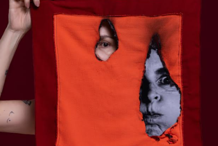 Imagem de uma pessoa segurando em lenço de cor laranja e vermelho, com um buraco no meio e um olho no meio