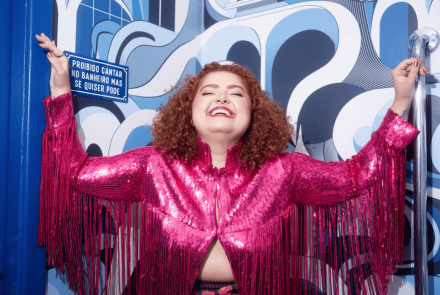 Mulher branca de cabelos cacheados e ruivos sorri com a cabeça para trás, de braços abertos, vestindo uma jaqueta rosa de paetês com franjas. Ao fundo, uma parede com estampa abstrata azul e branca exibe uma placa com os dizeres: "Proibido cantar no banheiro mas se quiser pode".