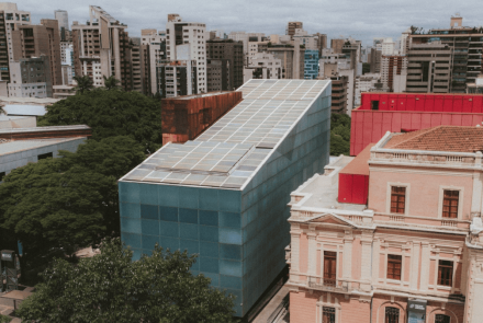 Fotografia aérea de um prédio moderno de formato inclinado com fachada azul e teto coberto por painéis solares. Ele está situado entre edifícios históricos de arquitetura clássica e uma área arborizada, com o horizonte da cidade ao fundo.