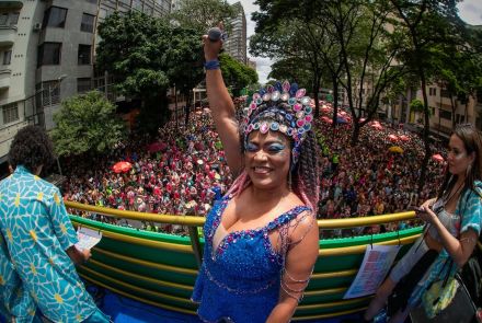 Três pessoas com fantasia de carnaval em cima do trio elétrico. Multidão ao fundo. Ensaio de Carnaval do Bloco Quando Come se Lambuza 2026