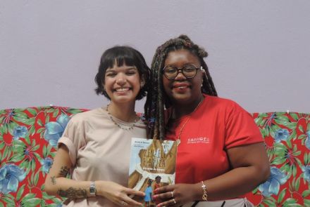 Duas mulheres ao centro segurando o livro "Zenzele e Aziza: através do tempo". Mulher branca à esquerda e mulher preta à direita.