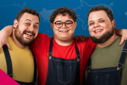 Três homens jovens e sorridentes aparecem abraçados do peito para cima, transmitindo alegria e união. Todos vestem macacões jeans sobre camisetas coloridas: o da esquerda veste amarelo, o do centro (que usa óculos redondos) veste vermelho, e o da direita veste verde-oliva. O fundo é azul com silhuetas sutis de porquinhos e um detalhe geométrico rosa no canto inferior.