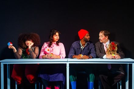 Quatro atores sentados atrás de uma mesa, imagem com fundo preto. Duas mulheres à esquerda e dois homens à direita. As mulheres vestem blusa longa e saia, os homens usam ternos e acessórios coloridos. 