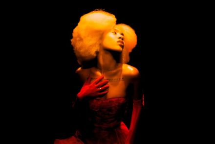 Mulher com cabelo volumoso e claro posa em fundo totalmente preto. Iluminada por luz quente, veste um vestido vermelho e luvas longas, com uma das mãos sobre o peito e o rosto inclinado para cima, criando uma cena dramática e teatral.