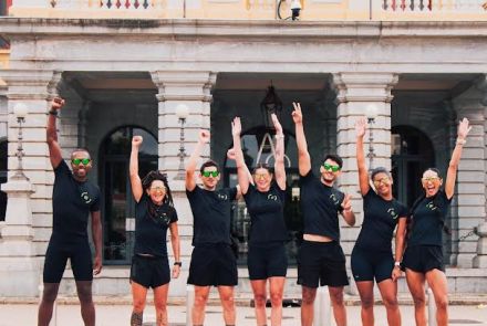 Sete atletas corredores em frente à Praça Rui Barbosa, há três homens e quatro mulheres. Eles vestem blusa e bermuda pretas e erguem um braço em comemoração.