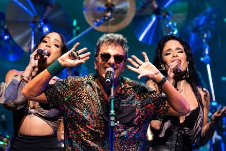 Cantor ao centro acompanhado de duas mulheres, uma em cada lado, reproduzindo vocal de apoio. Eles vestem roupas escuras com estampa, há instrumentos musicais ao fundo.