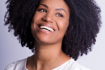 Artista Vavá Fernandes. Mulher preta sorrindo, veste blusa branca. Foto colorida sob fundo claro.