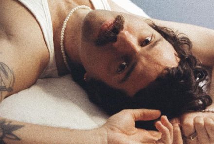 Artista Rafa Castro deitado com os braços próximos à cabeça enquanto encara a câmera com semblante sério. Veste camisa regata branca.