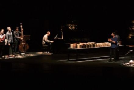 Cinco artistas no palco tocando instrumentos. Palco escuro com iluminação concetrada nos artistas.