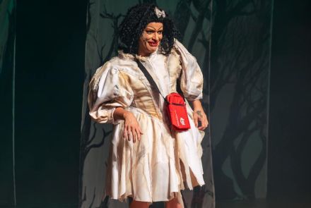 Suzy Brasil usando vestido branco com detalhes em amarelo, bolsa vermelha sobre os ombros e laço prata na cabeça. Imagem com fundo escuro.