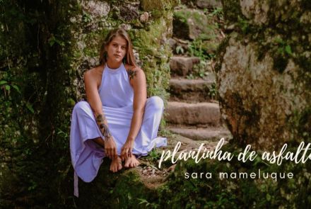 Artista Sara Mameluque sentada em um chão de pedra coberto por vegetação rasteira. Ela usa vestido lilás e os pés descalõ, encara a câmera com semblante sério.