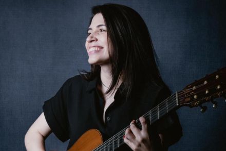 Artista Marina Aquino vestindo blusa de botão preta e segurando um violão enquanto sorri. Imagem com fundo escuro.
