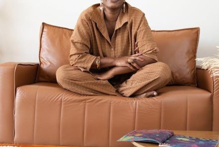 Arista Éle Fernandes (mulher preta) sentada de pernas cruzadas em um sofá marrom, ela sorri para a câmera. Exemplares do livro "Dinare" espalhados por uma mesa rendonda baixa e em cima do carpete claro. 