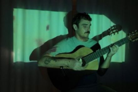 Artista Lucas Caram sentado tocando violão. Iluminação da foto direcionada ao artista.
