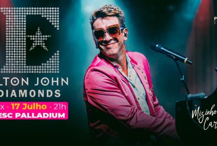 Artista Mizinha Carvalho. Homem sorridente, usando óculos chamativos e blazer rosa, toca piano e canta ao microfone sob luzes de palco.