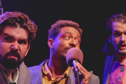 Três atores no palco usando terno e com expressões faciais de seriedade.