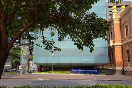 Foto do prédio do Espaço do Conhecimento UFMG. Edifício com fachada de vidro em tom azul claro, nome do local escrito em placa retangular fixa ao chão.