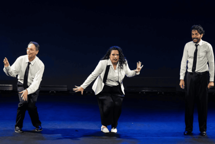 Três artistas — dois homens e uma mulher — realizam uma performance em um palco com fundo escuro e iluminação azulada. Todos vestem calças pretas e camisas brancas com gravatas. Eles exibem expressões dinâmicas e poses coreografadas, sugerindo um momento de teatro, dança ou improviso.