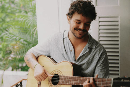 A imagem, com tons suaves e naturais, mostra o músico sorrindo levemente enquanto olha para as cordas de um violão acústico. Ele veste uma camisa listrada de manga curta e está posicionado em frente a uma janela com venezianas brancas abertas. Ao fundo, à esquerda, é possível ver a folhagem verde de palmeiras, criando uma ambientação tranquila e iluminada pela luz do dia.