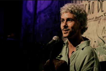 A imagem mostra o artista de perfil parcial, sorrindo suavemente enquanto canta em um microfone e toca violão. Ele tem cabelos curtos, cacheados e descoloridos, e veste uma camisa de tecido leve na cor verde-água. O fundo é escuro, com uma iluminação dramática que destaca tons de roxo à esquerda e uma parede com pichações e frases artísticas à direita, criando uma estética urbana e acolhedora de show ao vivo.