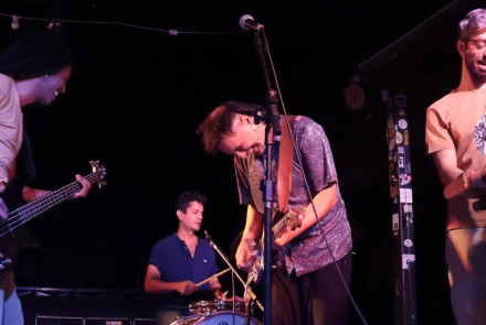 A imagem captura quatro músicos em um palco com iluminação focada e fundo escuro. No centro, o guitarrista — com uma camisa brilhante prateada — aparece inclinado sobre seu instrumento em um momento de entrega. À esquerda, um baixista sorri enquanto toca; ao fundo, o baterista mantém o ritmo concentrado; e à direita, o tecladista acompanha a performance com uma partitura à frente. O clima é de descontração e sintonia entre os artistas durante o show.