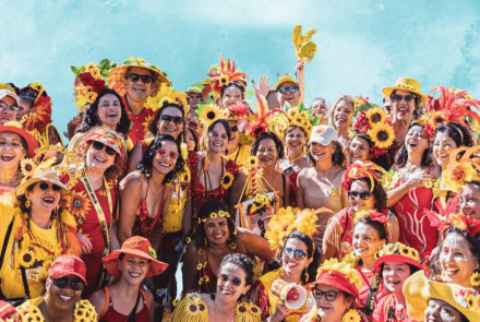 A imagem mostra um grande grupo de foliões sorridentes, a maioria mulheres, celebrando o Carnaval. Eles vestem fantasias vibrantes nas cores vermelho e amarelo, com destaque para diversos adereços de girassóis em chapéus, tiaras e roupas. A atmosfera é de alegria e união, com várias pessoas acenando para a câmera sob um céu azul claro ao fundo. É uma representação típica da energia festiva dos blocos de rua mineiros.