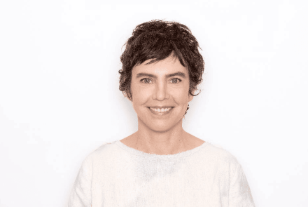 A imagem apresenta um plano médio centralizado da artista sorrindo de forma leve e acolhedora, contra um fundo totalmente branco e infinito. Ela possui cabelos escuros, curtos e levemente repicados, e veste uma blusa branca de tecido simples e textura natural. A iluminação é suave e frontal, destacando seus traços com clareza e transmitindo uma sensação de serenidade e minimalismo.