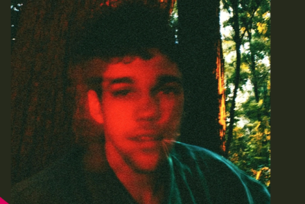 Fotografia em estilo granulado de um jovem de pele clara e cabelos escuros, com um efeito de dupla exposição que deixa seu rosto avermelhado. Ele está em uma floresta, com troncos de árvores e folhagens ao fundo iluminados pela luz do sol.