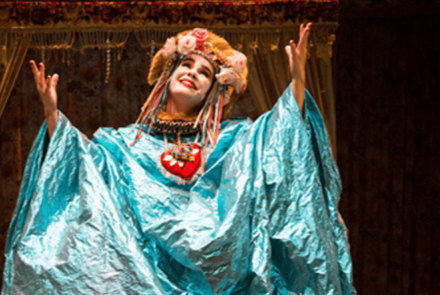 Imagem horizontal mostra uma atriz no palco, do busto para cima, caracterizada com um figurino teatral exuberante. Ela é uma mulher branca, com o rosto pintado de branco e bochechas rosadas, que sorri olhando para o alto com os braços abertos e as mãos erguidas. Veste uma túnica larga e brilhante em tom azul-celeste, um colar com um grande coração vermelho decorado no centro do peito e um adereço de cabeça dourado adornado com flores e fitas coloridas pendentes.